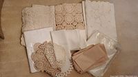 Overview of folded vintage beige lace doilies, white linens, and some tan folded linens on floor.