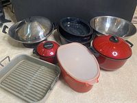 Photo showing steel wok with lid, three red metal pots with lids, black speckled enamel roasters, large stainless steel bowl, rectangular metal roasting pan with rack, and a red plastic Tupperware container.