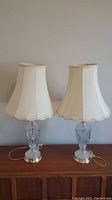 Full view of two matching lamps with glass bases, brass bottoms, and cream-colored scalloped fabric shades on a wooden sideboard.