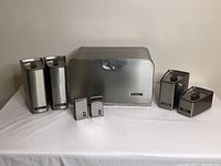 Wide front view of Lincoln Beauty Ware metal breadbox, flour and sugar canisters, tea and coffee canisters, and salt and pepper shakers arranged on white cloth.
