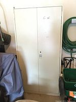 Front view of the tall off-white metal storage cabinet with two doors, showing minor scuff marks and the cabinet standing in a garage setting.