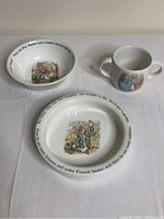 Set of three child dish items including large bowl, small bowl, and two-handled mug, all white ceramic with Peter Rabbit illustrations and printed story text around rims.