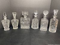 Six clear pinwheel pattern glass decanters arranged side-by-side against a black background, showcasing variety in stopper shape and bottle design.