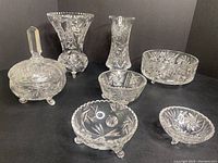 Seven clear crystal glass items arranged on black cloth showing detailed cut pinwheel and star patterns including two vases, three footed bowls, one covered candy dish, and one larger footed bowl.