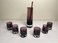Overview of full set: pitcher with stirring rod surrounded by six tumblers on white surface.
