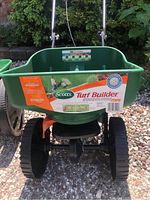 Front view of a Scotts Turf Builder EdgeGuard Mini lawn feeder showing green hopper, orange parts, and black wheels