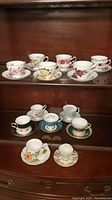 Photo showing 12 assorted china tea cups and matching saucers displayed on a wooden shelf in a display cabinet. Floral and traditional pattern styles are visible in various colors and detailing.
