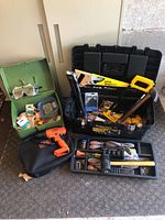 Overall view of the assorted tools including tool trays with hand tools, large black toolbox holding various tools, Stanley handsaw, rubber mallet, snake light, and the empty orange cordless drill case.