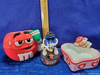 Photo of the three M&M's collectibles including red M&M ceramic figure, blue M&M animated saxophone player figurine, and heart-shaped dish with red M&M figure.