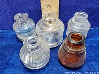 Five old glass ink bottles displayed on a blue cloth with a ruler for size reference. Four bottles are clear glass with varying shapes, and one bottle is amber glass.