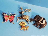 Photo of four vintage animal brooches on blue background showing colorful butterfly, gold-tone reindeer, small gold-tone dog, and black rhinestone elephant.