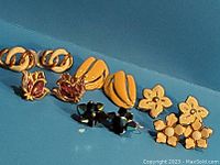 Close-up of assorted vintage earrings on blue background including gold-tone chain link, red floral enamel screw backs, black faceted studs, cream and gold clip-ons, and gold-tone floral pierced earrings.