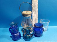 Five vintage glass items on blue background including three cobalt blue miniatures, one clear glass lantern with metal top, and one clear glass tea measure cup. A ruler is included for size reference.