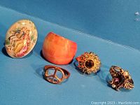 Five vintage costume rings displayed on blue background, including enamel oval nature scene, orange bangle, peace sign, sunflower with amber crystal, and ornate purple gemstone and pearls.