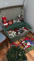 Photo of large green plastic tote bin filled with various Christmas decorations, including a musical Santa figure, a child figurine in red, a mailbox figure, and other small Christmas items.