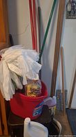 Photo showing red mop bucket with wringer, white disposable gloves inside, mop handles visible, and some cleaning bottles around