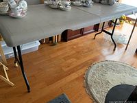 Full length side view of gray plastic top folding table with metal black folding legs and gold capped feet on a wood floor with tea set on top.