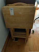 Four wooden folding tray tables stacked vertically within their rack on a hardwood floor, showing rectangular tabletops with light wood finish and rounded edges.