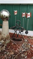 Photo showing full gazing ball on beige pedestal, rusty metal garden stand, and three Canadian flag solar stakes.