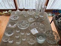 Photo of the full glassware collection displayed on a table near a window including vases, candy dishes, serving plates, dessert cups, and a bell.