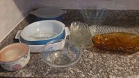 Overview of various glassware pieces on countertop including Pyrex blue handled bowl, amber glass dishes, clear glass baking dish, small floral patterned bowl, and covered casserole dishes