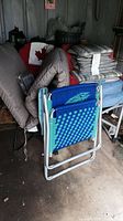 Photo showing two folding lawn chairs and stacked seat cushions in a garage or outdoor storage area. Left chair is a padded gray canvas style, center chair is blue woven plastic with metal frame, cushions stacked behind.