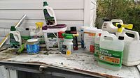 Photo of multiple lawn chemical containers on old white table, showing jugs, spray bottles, and smaller bottles with partial contents visible.