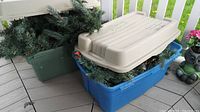 Photo showing two plastic storage totes, one green and one blue, both filled with faux green garland and string lights, with a beige lid resting on top of the blue tote.