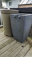 Two rain barrels side by side on wooden deck, square gray barrel in front and round beige barrel behind