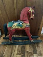 Full side view of the ornate wooden rocking horse showing red paint and decorative gold and green saddle and harness details.