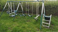 Full side view of the swing set showing two plastic swing seats, a glider bench swing, teeter-totter, and trapeze bar attached to a metal frame in a grassy backyard.