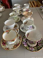 Full view showing ten assorted tea cups and saucers arranged in two rows, featuring multiple floral patterns and gold trim