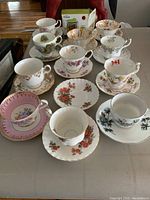 Eleven assorted fine china tea cups and saucers arranged on table plus boxed Trudeau mug with infuser in background.