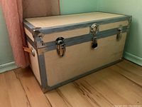 Oblique front view of light-colored wooden trunk with metal edges and lock, sitting on wooden floor next to a pink curtain.