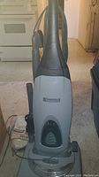 Front view of gray Kenmore upright vacuum cleaner showing power cord wrapped around base and visible vacuum bags nearby.