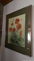 Framed watercolor painting of red-orange poppies showing the full piece hung on a wall from an angled side view.