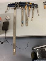 Photo showing six metal bar clamps of varying lengths and handle types laid out on a table, longest reaching up to 3 feet.