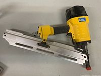 Side view of yellow and black pneumatic framing nailer with metal rail loaded with nails, resting on a light surface.