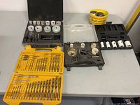Full view of three hole saw kits, a plastic bin with hole saw bits, and a yellow drill bit case on a table