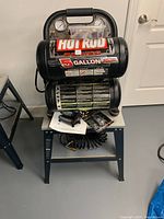 Front view of the black Hot Rod 5 Gallon twin tank air compressor mounted on metal stand, with pressure gauges and accessories on the stand shelf.