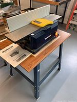 Blue Mastercraft jointer/planer on metal stand with wood top, yellow push block on table, operation manual visible
