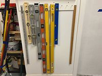 Photo showing seven different spirit levels of varying lengths and colors hung vertically on a white wall. Two wooden rulers are also hung vertically next to the levels.