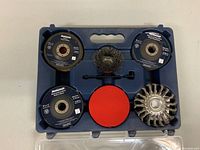 Angle grinder accessory wheels and discs arranged in blue carrying case, showing black flap discs, metallic twisted wire wheel, red backing pad, and other grinding accessories.
