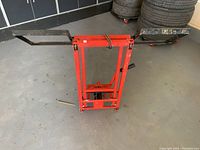 Front view of the Pro-Lift lawn mower lift showing red frame, black lifting arms spread open, and foot pedal mechanism on the floor.