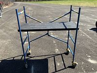 Front and side views of blue metal scaffold with LITE branded black board and caster wheels on pavement outdoors.