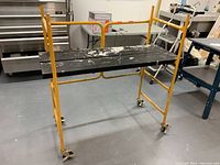 Front side view of mobile scaffold bench showing yellow tubular metal frame, black wooden planks, and caster wheels.