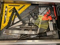 Top-down photo showing mixed measuring tools including Stanley yellow square ruler, black triangular rulers, metal calipers, protractor, and a silver staple gun in a drawer.
