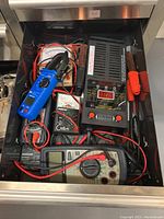 Top-down photo of metal drawer holding assorted electrical testing and measurement tools including multimeters, clamp meter, test leads and battery tester.