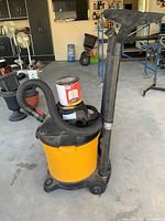 Front angled view showing the vacuum's yellow body, hose coiled to one side, and the wide nozzle attachment held vertically on the back.