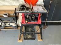 Front view of red Skilsaw table saw on black metal stand showing control knobs, power switch, rip fence, safety guard, and user manual on table surface.